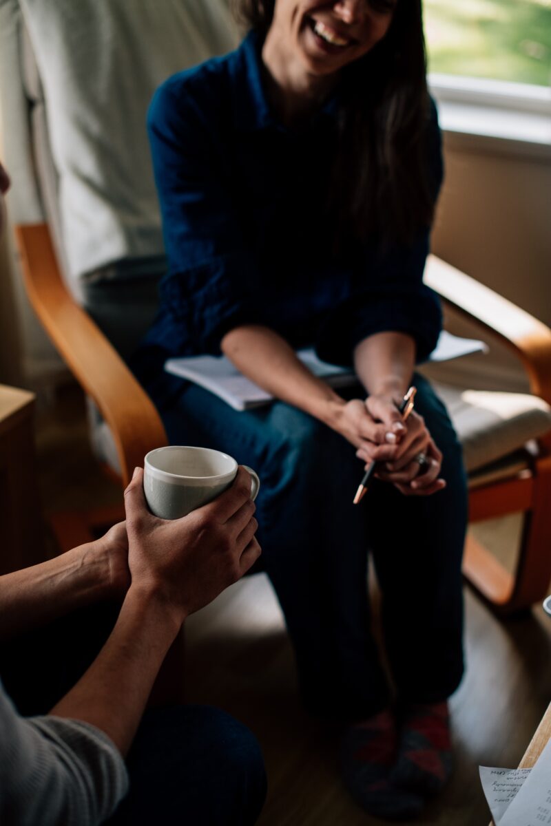 A smiling woman sat down holding a pen, next to a colleague holding coffee. They contemplate Millennials vs Gen X in the Workplace!