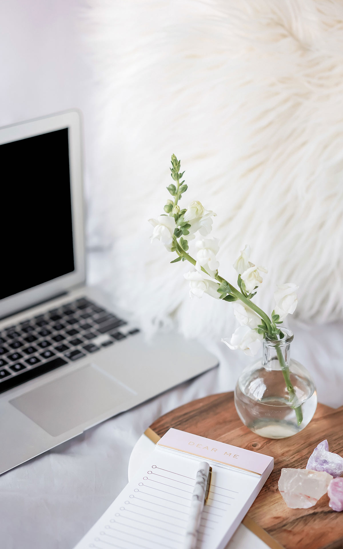 macbook on bed, side table with flower vase, notebook and crystals.