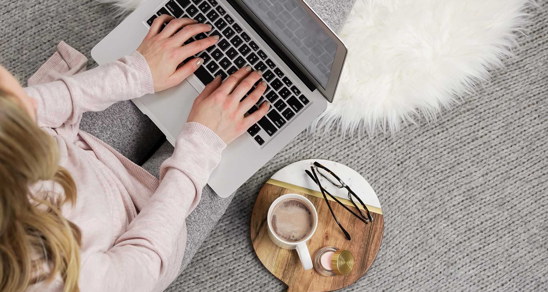 woman typing on laptop with coffee and glasses on side table.